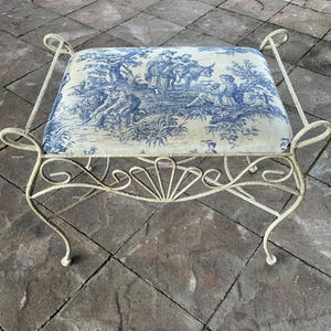May include: A decorative bench with a cream-colored, wrought-iron frame. The seat cushion features a blue and white toile pattern depicting a pastoral scene. The bench has curved legs and decorative scrollwork.
