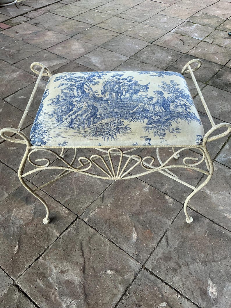 May include: A decorative bench with a cream-colored, wrought-iron frame. The seat cushion features a blue and white toile pattern depicting a pastoral scene. The bench has curved legs and decorative scrollwork.