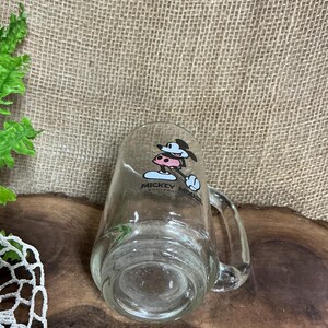 May include: Clear glass mug featuring Mickey Mouse in a cowboy hat. The mug has a handle and a printed design of Mickey Mouse. The mug is transparent, allowing the contents to be visible. The mug is a collectible item.