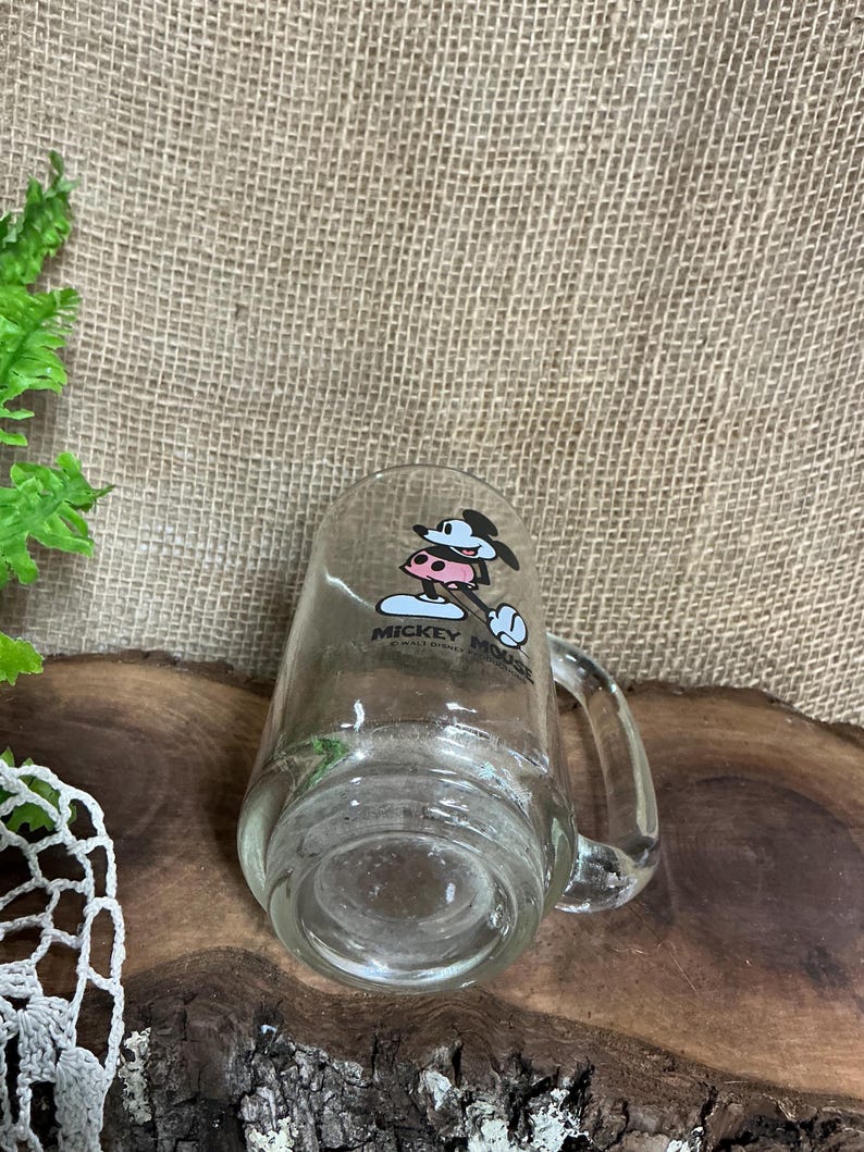 May include: Clear glass mug featuring Mickey Mouse in a cowboy hat. The mug has a handle and a printed design of Mickey Mouse. The mug is transparent, allowing the contents to be visible. The mug is a collectible item.