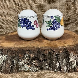 May include: A pair of white ceramic salt and pepper shakers. Each shaker features a fruit design with grapes, cherries, and a lemon. The shakers have a decorative pattern around the top and are placed on a wooden surface.
