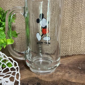 May include: Clear glass mug featuring a classic Mickey Mouse graphic in black, white, and red. The mug has a large handle and a wide base. The text "Mickey Mouse" is printed on the mug.