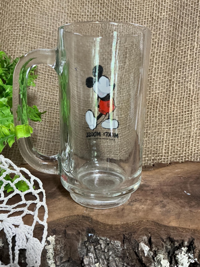 May include: Clear glass mug featuring a classic Mickey Mouse graphic in black, white, and red. The mug has a large handle and a wide base. The text "Mickey Mouse" is printed on the mug.