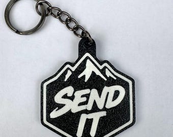 Puede incluir: Un llavero negro con superficie texturizada que presenta un gráfico de montaña en blanco y el texto "SEND IT" en una fuente negrita y estilizada. El llavero está unido a una anilla metálica circular mediante una cadena corta de metal.