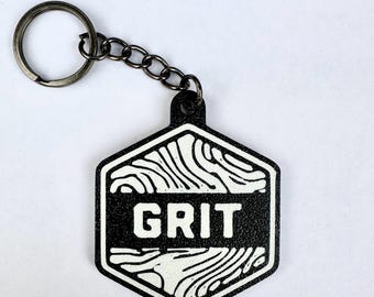 Puede incluir: Un llavero hexagonal con un diseño en blanco y negro con la palabra GRIT impresa en el centro. El diseño incluye patrones abstractos de vetas de madera blanca encima y debajo del texto, unido a una anilla y cadena de metal oscuro.