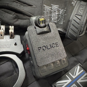 May include: A close-up shot of a black body camera with the word "POLICE" on it, a black glove, handcuffs, and a patch with a blue line and the Union Jack. The body camera has the text "REVEAL D-SERIES".