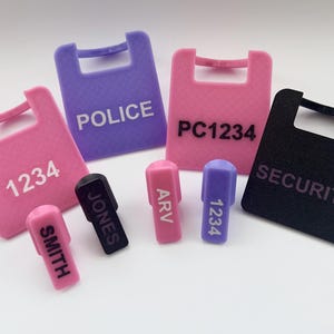 Puede incluir: Una colecci&oacute;n de etiquetas de seguridad y llaveros de colores. Las etiquetas son rosas, moradas y negras, con las palabras "POLICE", "SECURITY" y n&uacute;meros impresos. Los llaveros son rosas, morados y negros, con nombres y n&uacute;meros.