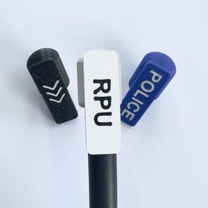 Puede incluir: Tres tapas de bol&iacute;grafo en negro, blanco y azul. La tapa negra tiene flechas blancas, la tapa blanca tiene las letras "RPU" y la tapa azul dice "POLICE" en blanco. Sobre un fondo blanco.