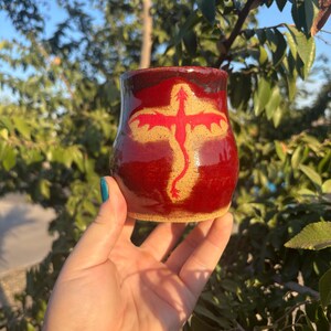 May include: A handmade ceramic mug with a deep red glaze. The mug features a stylized red dragon design on a tan background. The mug is held in a hand against a backdrop of green foliage.