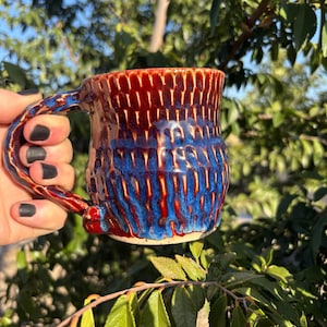 May include: Handmade ceramic mug with a textured surface. The mug features a vibrant color scheme of red and blue, with a glossy finish. The handle is integrated into the design. The mug is perfect for enjoying coffee or tea.