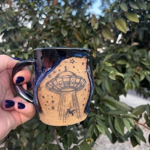 May include: A handmade ceramic mug with a black and blue glazed exterior. The mug features a detailed illustration of a UFO with a beam of light and a small figure. The mug is held against a backdrop of green foliage.