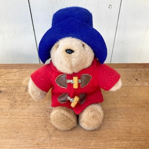 May include: A plush teddy bear wearing a bright blue hat and a red coat with wooden toggle closures. The bear has a tan face and paws, and is sitting on a wooden surface.