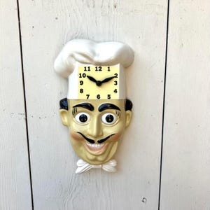 May include: A novelty wall clock shaped like a chef's head, featuring a smiling face with a black mustache and a white chef's hat. The clock face is square with black numbers and hands. The clock is mounted on a white wooden surface.