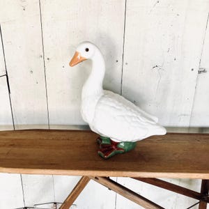 May include: A ceramic goose figurine featuring a glossy white body, a smooth orange beak, and dark brown eyes. The goose stands on a textured green base with dark red webbed feet, captured in a side profile view with its neck curved gracefully.