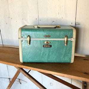 May include: A rectangular teal train case with a textured finish, featuring cream-colored trim, a top handle, and gold-tone metal latches. The front includes a central keyhole and a small rectangular plate with the letters PAS embossed in black.