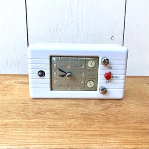 May include: A white rectangular General Electric clock with a central square face featuring black numerals and hands. The device includes two small circular dials labeled END and BEGIN, a red push button, and two silver knobs on the right side.