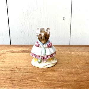 May include: A ceramic figurine of a mouse wearing a white dress with a pink and red checkered trim. The mouse has brown fur, a blue collar, and a white bonnet. The figurine is on a yellow base.