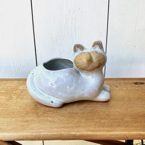 May include: A ceramic cat-shaped planter with a glossy, speckled off-white glaze. The cat figure is in a reclining position with its head turned forward, featuring a tan-colored face with etched details for the eyes, nose, and whiskers.