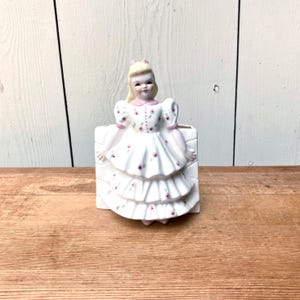 May include: A ceramic wall pocket planter shaped like a girl with blonde hair wearing a white dress with pink and green floral patterns. The dress features a tiered, ruffled skirt design, and the figure has painted facial features and a pink bow.