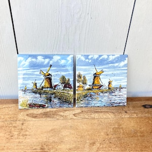 May include: Two square ceramic tiles featuring a hand-painted landscape scene of Dutch windmills, small houses, and a river with boats. The artwork uses a palette of blue, yellow, and green, depicting a cloudy sky over a rural waterfront setting.