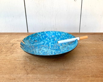 Könnte beinhalten: Ovaler Aschenbecher mit strukturierter blauer Oberfläche und einer Zigarette. Der Aschenbecher hat einen dunklen Rand und steht auf einer Holzoberfläche. Die blaue Farbe hat einen Marmoreffekt. Mit einer Kerbe für die Zigarette.