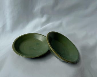 May include: Two small, round, olive-green ceramic dishes. The dishes have a slightly raised edge and a smooth, glossy finish. The dishes are displayed on a white background.