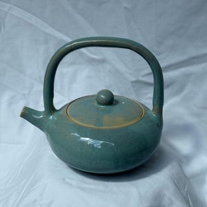 May include: A round ceramic teapot with a glossy teal glaze and a high, arched handle. The teapot features a matching lid with a small knob handle and a short, pointed spout. The rim of the lid and the base of the handle show subtle tan accents.