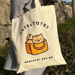 May include: Cream tote bag with the text "CATS & TOTES" and "wherever you go." The bag features a cartoon illustration of an orange tabby cat in an orange carrier. The bag has cream-colored handles.