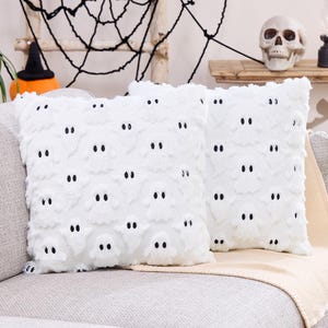 May include: Two white decorative cushions with a ghost pattern. Each cushion features a repeating design of ghost faces with black eyes. The cushions are placed on a grey sofa, with Halloween decorations in the background.