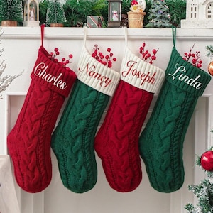 May include: Four cable-knit Christmas stockings in red and green hang from a white mantel. Each stocking is personalized with a name embroidered in white: Charles, Nancy, Joseph, and Linda. The stockings are decorated with red berries.