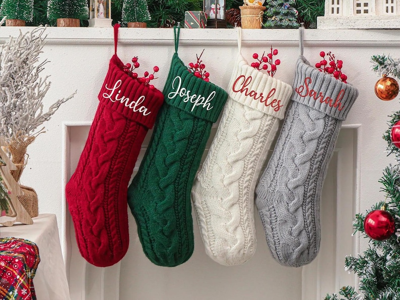 May include: Four cable-knit Christmas stockings in red, green, cream, and gray hang from a mantel. Each stocking is personalized with a name in a complementary color: "Linda", "Joseph", "Charles", and "Sarah". Red berries adorn the top of each stocking.