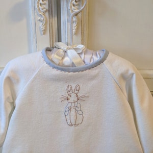 May include: White baby onesie with a blue trim around the neckline. The onesie has an embroidered image of Peter Rabbit in brown and blue.