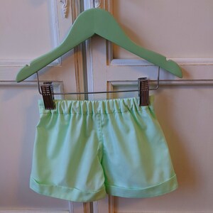 May include: Light green shorts with a cuffed hem, hanging on a green wooden hanger.