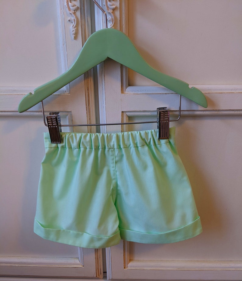 May include: Light green shorts with a cuffed hem, hanging on a green wooden hanger.
