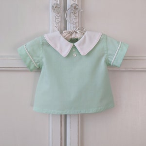 May include: A light green short-sleeved shirt with a white collar and white trim around the sleeves. The shirt has a button closure at the neck.