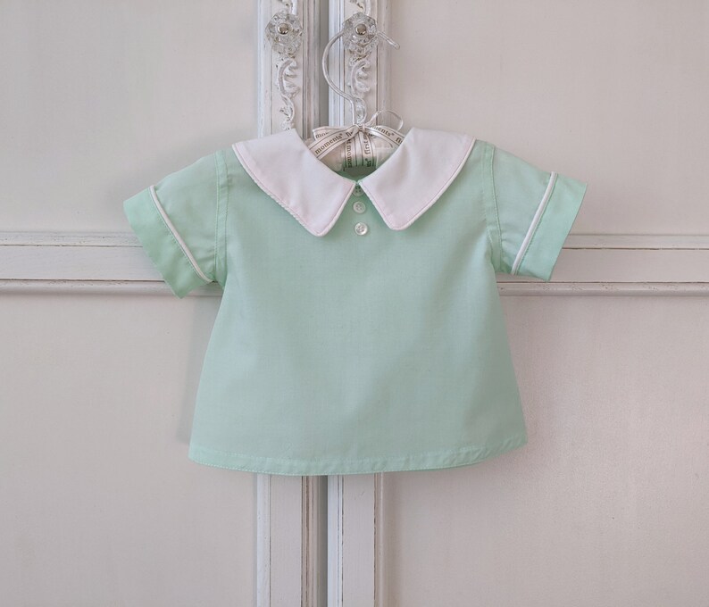 May include: A light green short-sleeved shirt with a white collar and white trim around the sleeves. The shirt has a button closure at the neck.