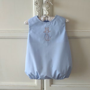 May include: A pale blue baby bubble romper featuring a Peter Rabbit embroidery. The sleeveless romper has a rounded neckline and a gathered hem. It is displayed on a white hanger against a white backdrop.