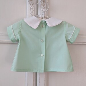 May include: A light green short-sleeved shirt with a white collar and white trim around the sleeves. The shirt has a button-up front.