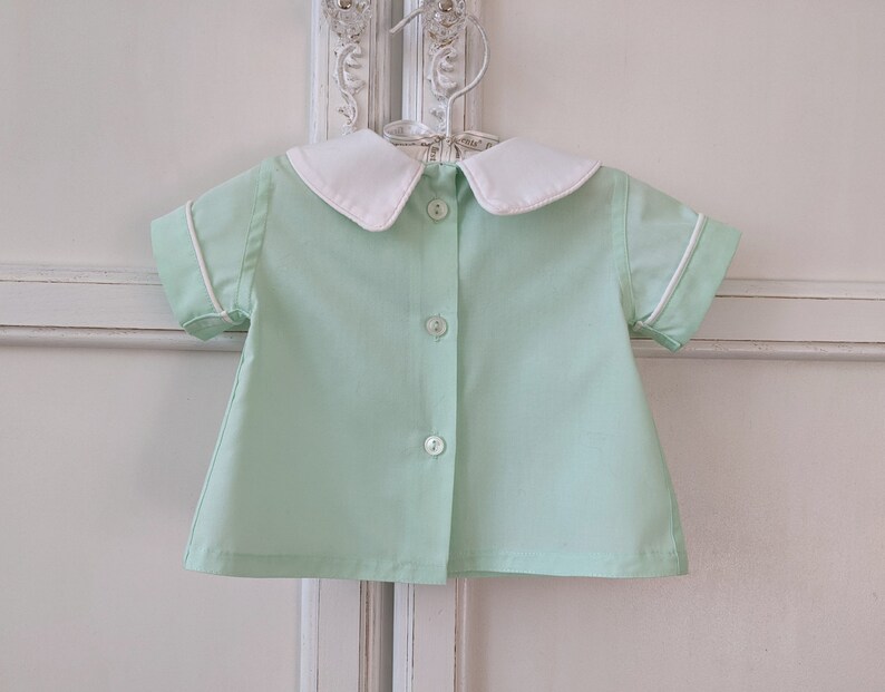 May include: A light green short-sleeved shirt with a white collar and white trim around the sleeves. The shirt has a button-up front.