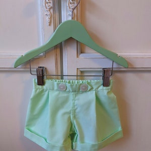 May include: Light green shorts with a button closure and a cuffed hem. The shorts are hanging on a light green wooden hanger.