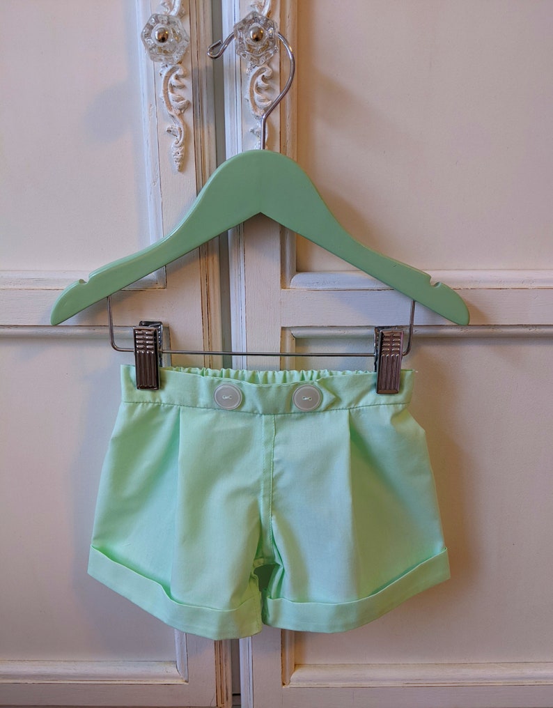 May include: Light green shorts with a button closure and a cuffed hem. The shorts are hanging on a light green wooden hanger.