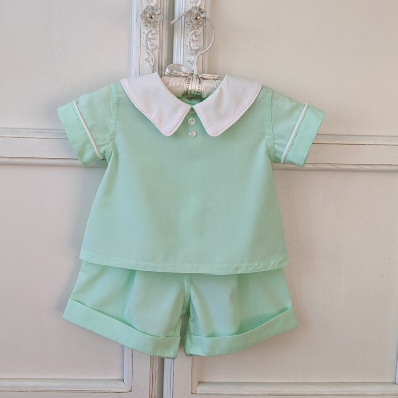 May include: A light green two-piece outfit for a baby or toddler. The outfit includes a short-sleeved shirt with a white collar and a pair of shorts.