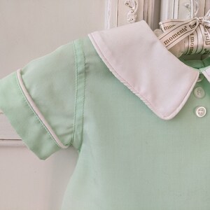 May include: A light green baby shirt with a white collar and white trim around the sleeves. The shirt has two white buttons on the front.