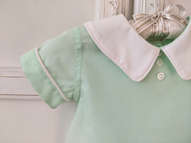 May include: A light green baby shirt with a white collar and white trim around the sleeves. The shirt has two white buttons on the front.