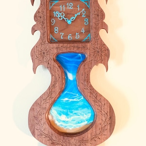 May include: A wooden wall clock with a carved design featuring a vase with a blue and white resin ocean scene. The clock face has Roman numerals and a blue border.