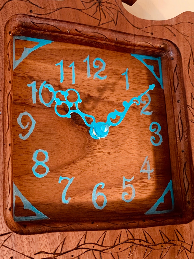 May include: A wooden clock face with blue painted Roman numerals and hands. The clock face is set in a wooden frame with carved details.