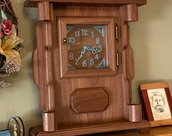 May include: A wooden clock with a glass face and a decorative design. The clock face has Roman numerals and a blue and white design. The clock is sitting on a wooden shelf.
