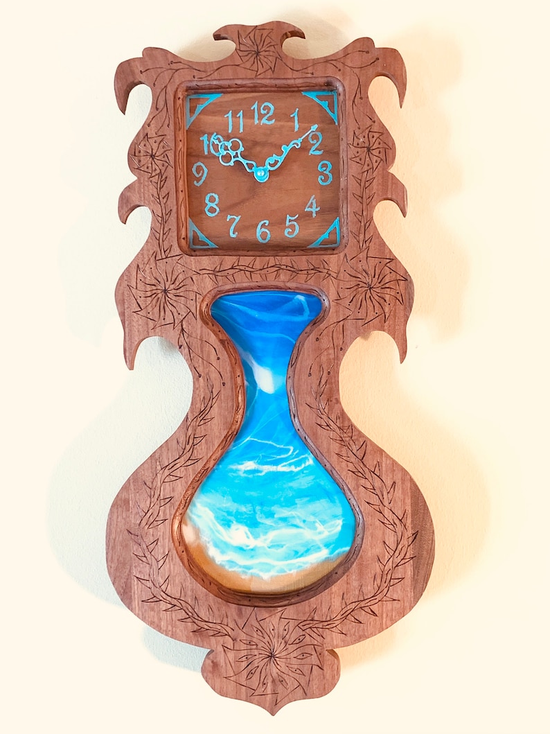May include: A wooden wall clock with a carved design featuring a vase with a blue and white resin ocean scene. The clock face has Roman numerals and a blue border.