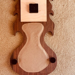 May include: A wooden clock face with a decorative design. The clock face is made of walnut wood and has a brown finish. The clock face is attached to a wooden backing that is also made of walnut wood. The clock face is covered with a piece of brown paper. The clock face has two black metal knobs on the bottom.