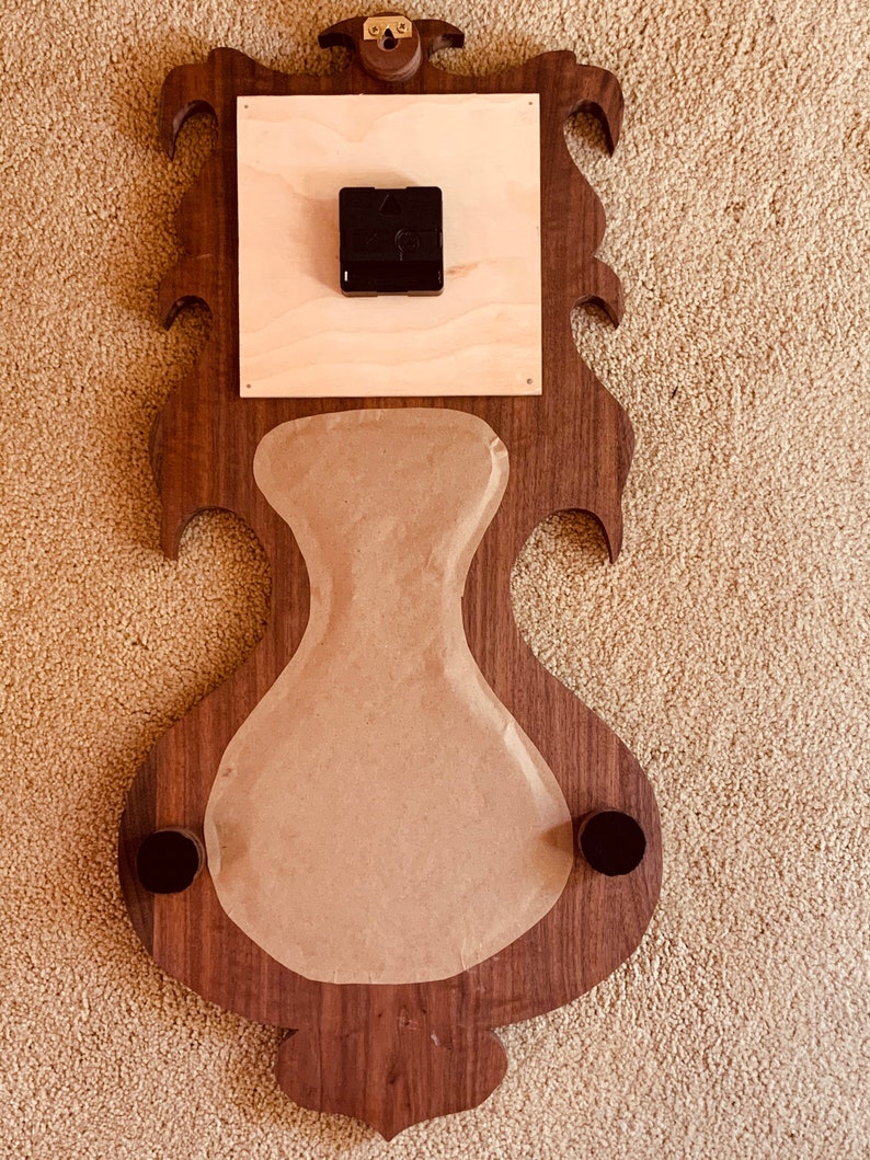 May include: A wooden clock face with a decorative design. The clock face is made of walnut wood and has a brown finish. The clock face is attached to a wooden backing that is also made of walnut wood. The clock face is covered with a piece of brown paper. The clock face has two black metal knobs on the bottom.